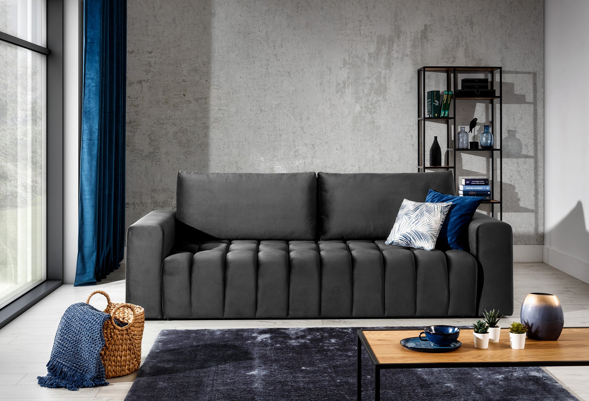 Sofa Lazaro Monolith 97 ciemnoszary w stylowym salonie przy dużym oknie, obok plecionego kosza z pledem i metalowej regału z dekoracjami na tle betonu i granatowych zasłon.