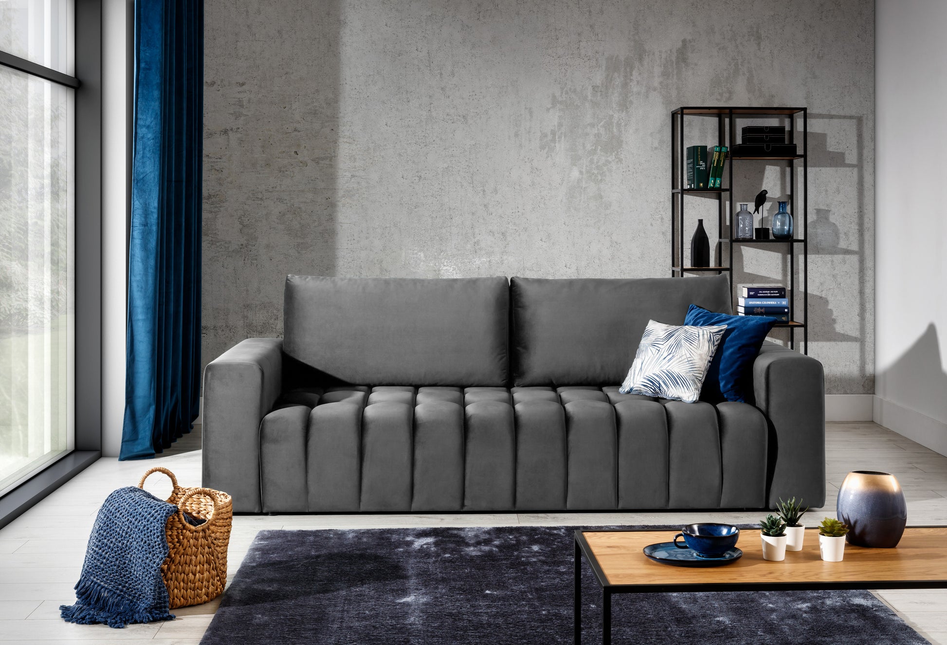 ofa Lazaro Nube 05 ciemnoszara w stylowym wnętrzu: sofa ustawiona przy surowej betonowej ścianie, obok dużego okna z granatowymi zasłonami i wiklinowego kosza, na tle stolika kawowego i regału, tworząca loftową aranżację.