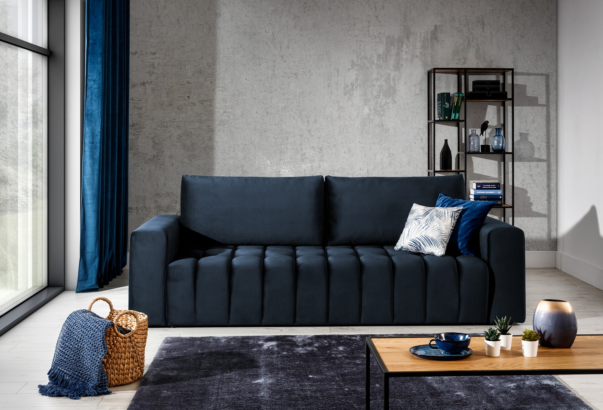 Sofa Lazaro Monolith 79 granatowa w stylowym wnętrzu: sofa ustawiona przy betonowej ścianie obok dużego okna z granatowymi zasłonami, uzupełniona metalowym regałem, wiklinowym koszem i stolikiem kawowym, tworząc nowoczesną, loftową aranżację.