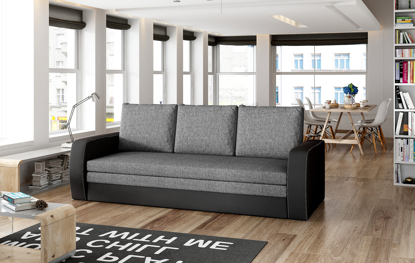 Sofa Inversa w odcieniach Sawana 05 ciemnoszary i Soft 11 czarny ustawiona w nowoczesnym, jasnym salonie z jadalnią w tle.