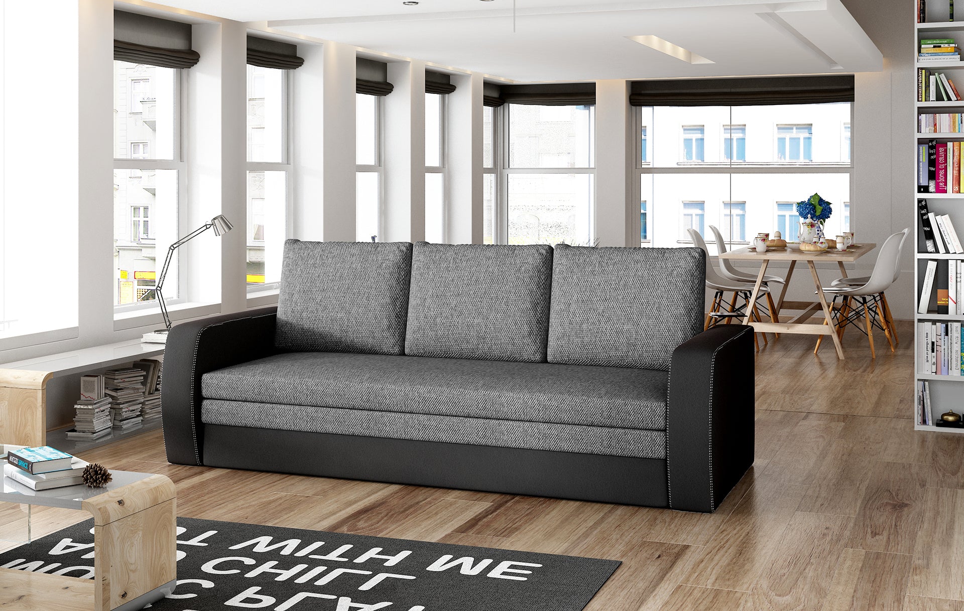 Sofa Inversa w odcieniach Sawana 05 ciemnoszary i Soft 11 czarny ustawiona w nowoczesnym, jasnym salonie z jadalnią w tle.