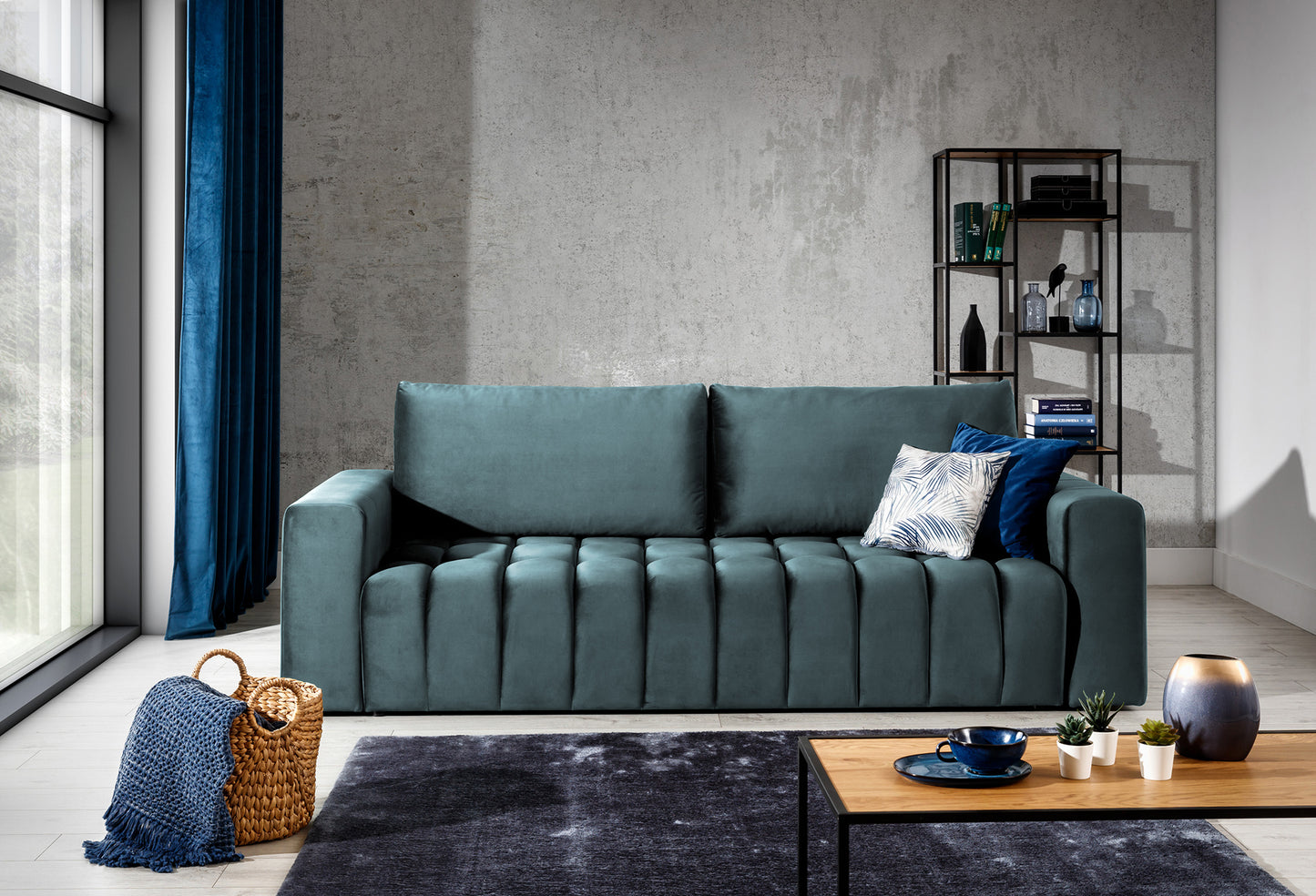Sofa Lazaro Monolith 76 jasnoniebieska w aranżacji salonu: sofa ustawiona na tle betonowej ściany obok dużego okna z granatowymi zasłonami, uzupełniona metalowym regałem, wiklinowym koszem z kocem i stolikiem kawowym, podkreślając nowoczesny charakter wnętrza.