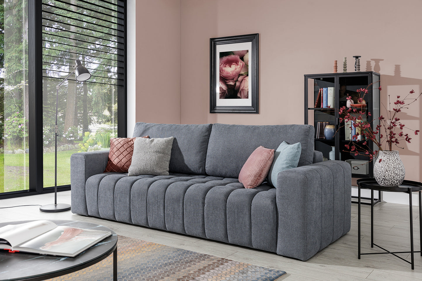 Sofa Lazaro Primo 89 w aranżacji salonu: jasnoszara tapicerka, solidne podłokietniki i przeszywane siedzisko w pomieszczeniu z różowawą ścianą, przeszklonym oknem i stolikiem kawowym.