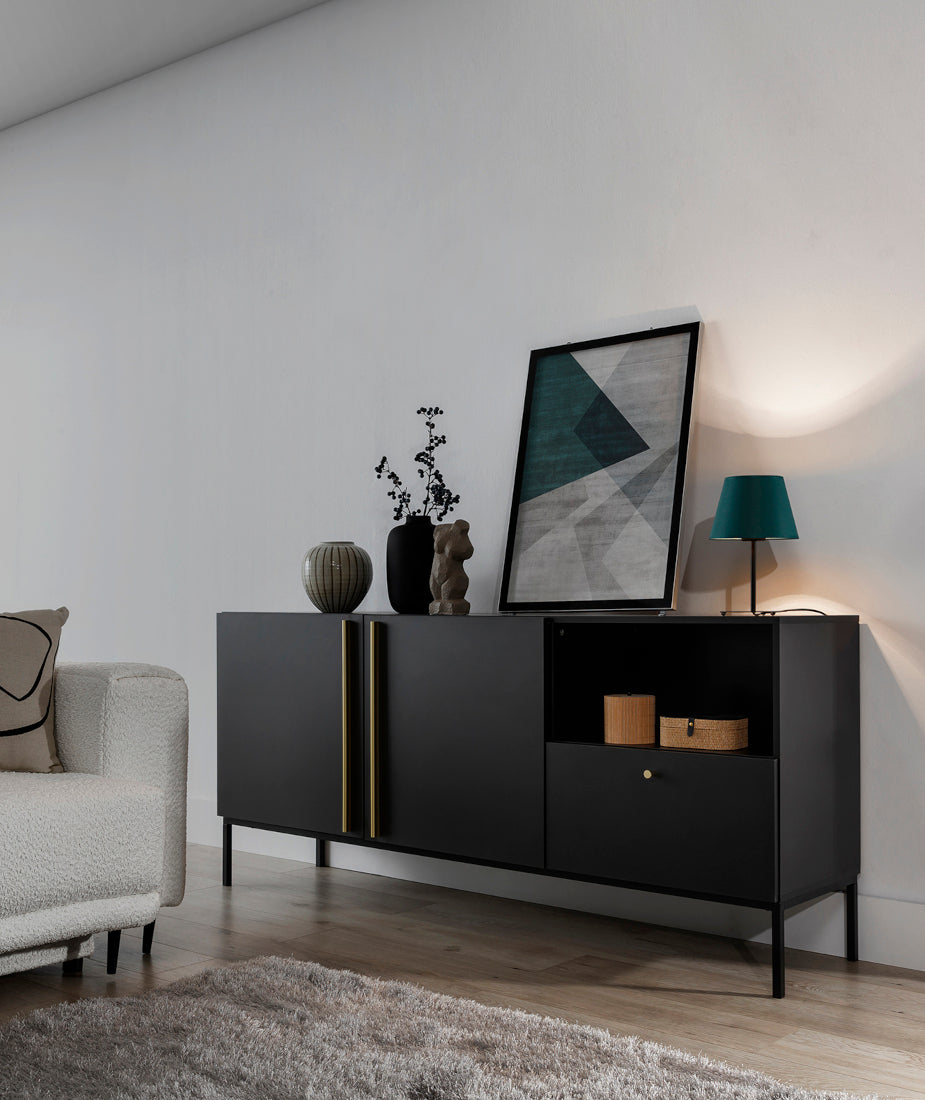 Włączona lampka nocna Vespillo 2a z zielonym abażurem i złotym wnętrzem, rzucająca ciepłe światło na białą ścianę i czarną komodę, tworząca przytulny klimat.