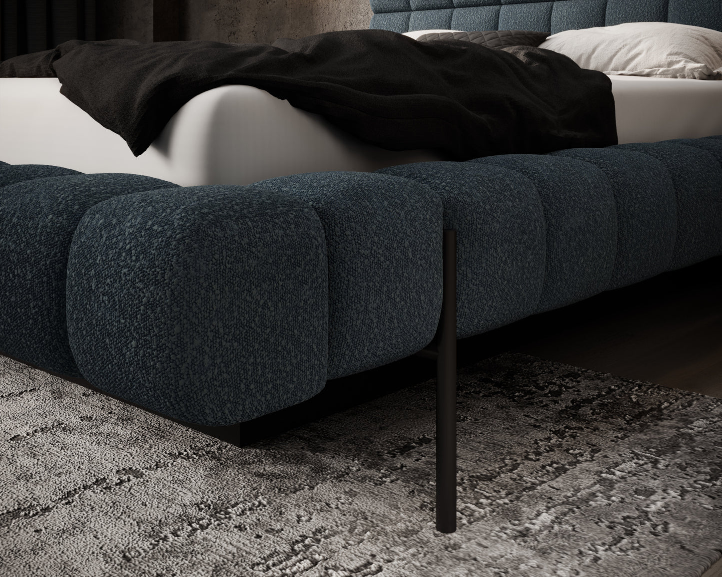 Zbliżenie na narożnik łóżka Magentta w tkaninie Jaffray 40 niebieski, z czarną metalową nogą na tle miękkiego dywanu.