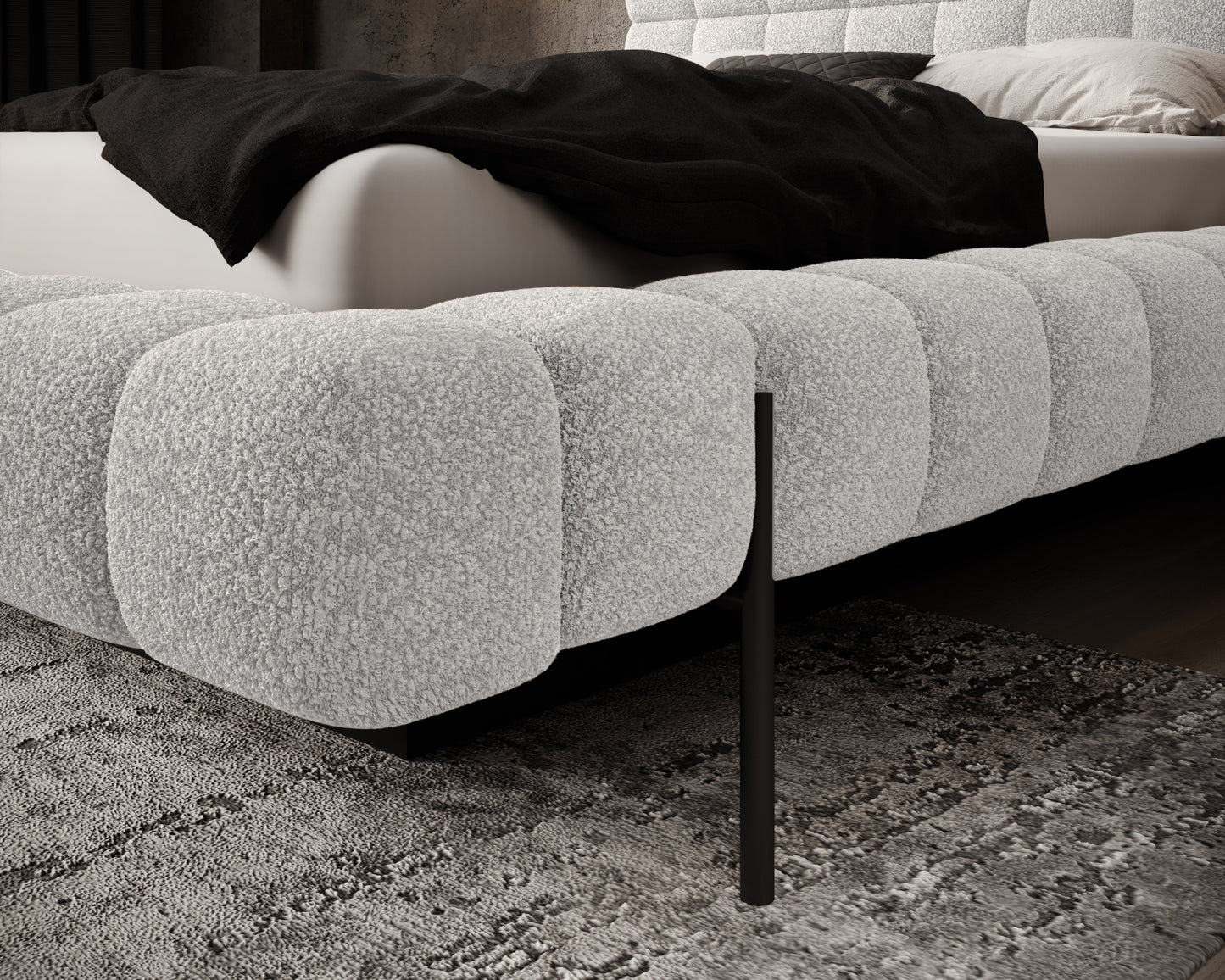 Zbliżenie na narożnik łóżka Magentta w tkaninie Royal 01 biały – zaokrąglona forma i czarna metalowa nóżka na tle dywanu.