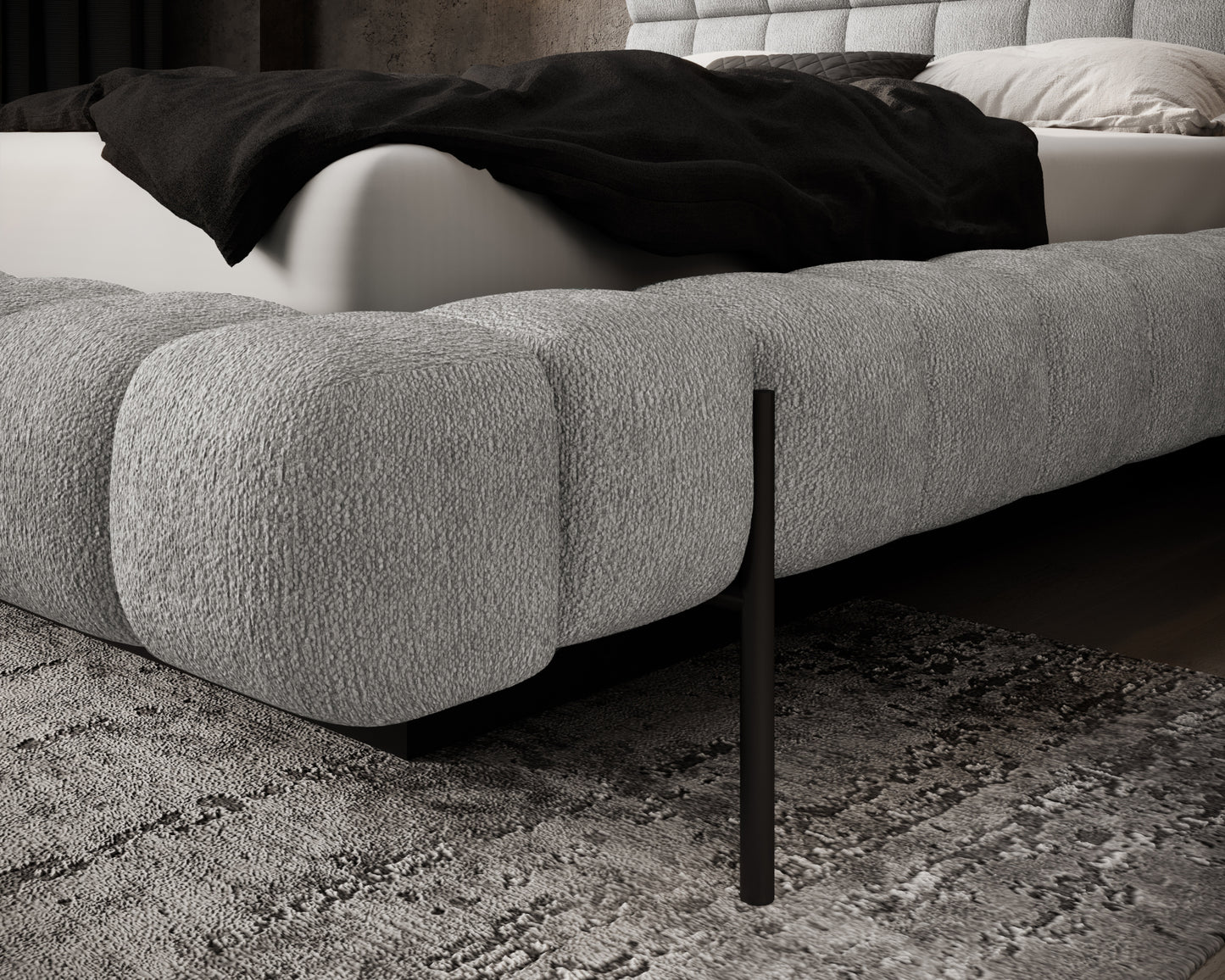 Boczne ujęcie łóżka Magentta z pikowanym bokiem w tkaninie Touch 02 jasnoszary i czarną nogą na tle dywanu o grubym splocie.