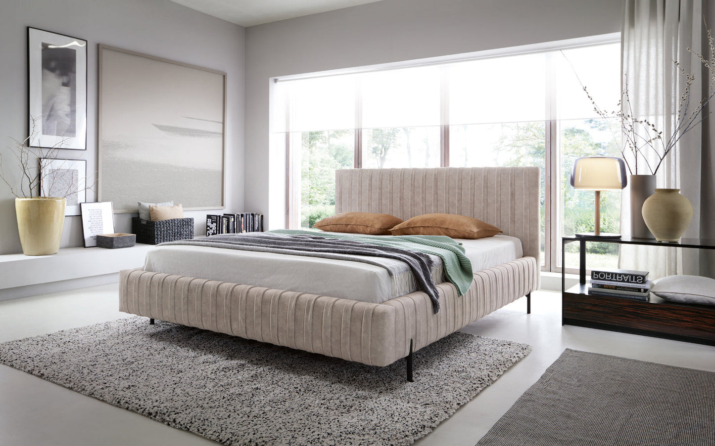 Łóżko Plissa 160×200 w kolorze Relax 18 beżowy ustawione na wzorzystym dywanie w nowoczesnej sypialni przed dużym oknem, ozdobione narzutą i poduszkami.