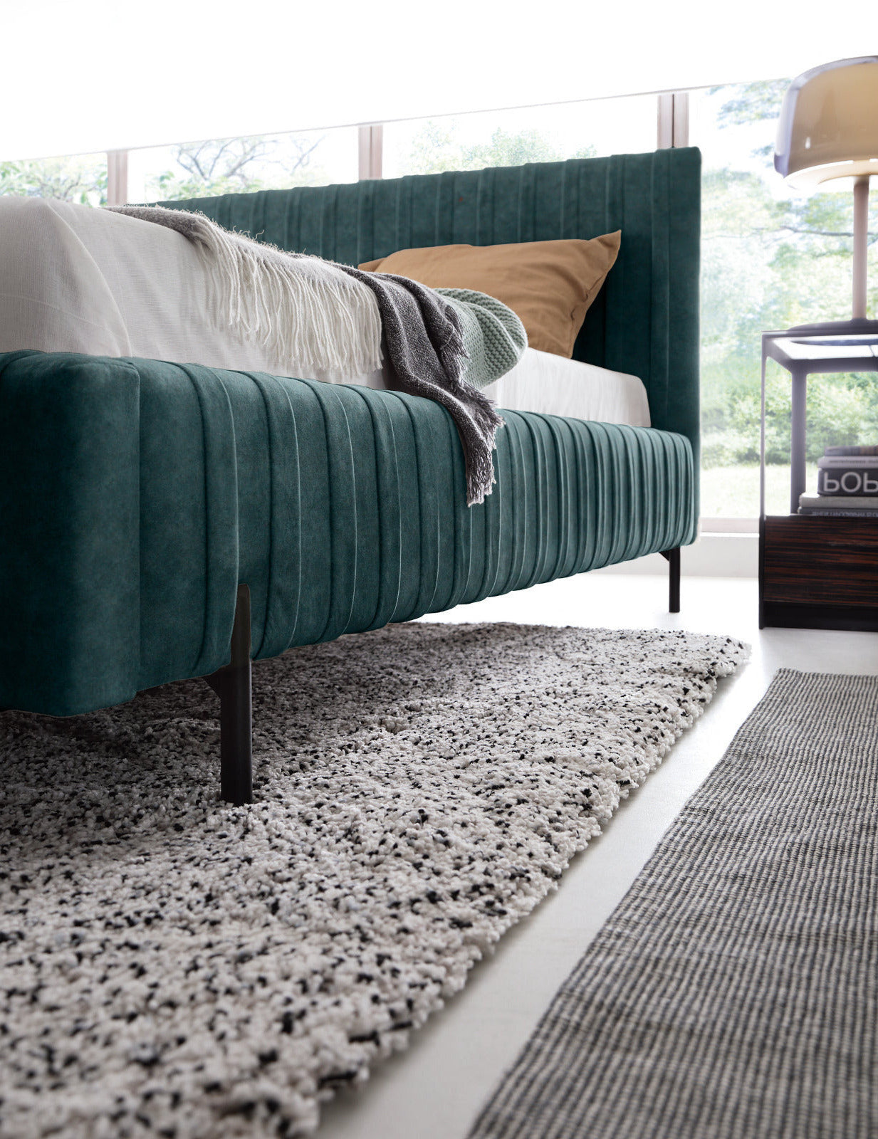 Niski kąt patrzenia na bok i nogę łóżka Plissa 180×200 w kolorze Relax 38 zielony, stojące na jasnym dywanie obok stolika.