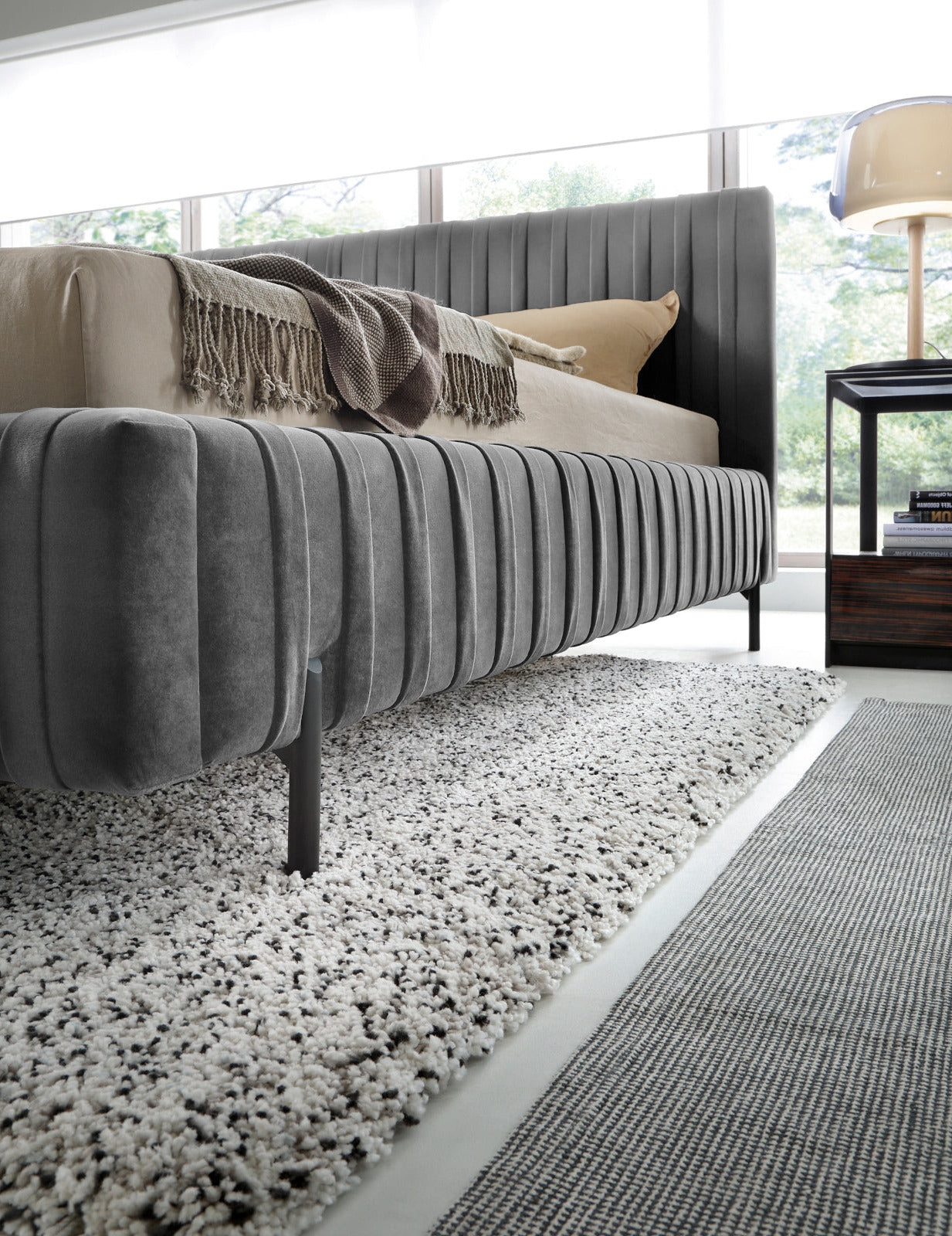 Perspektywa boczna łóżka Plissa 140×200 Velvetmat 04 szare nad puszystym dywanem, eksponująca charakterystyczną ramę i nóżki.