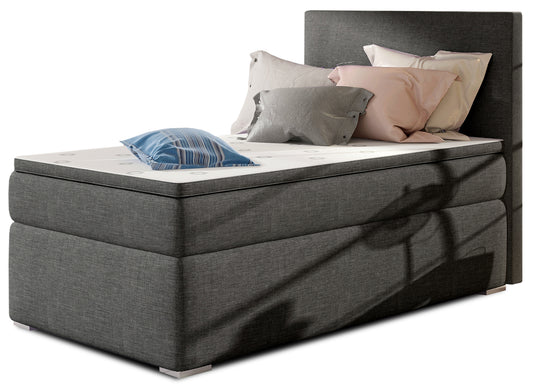 Łóżko kontynentalne Rocco w kolorze Sawana 05 ciemnoszary, układ lewy – widok studyjny z przodu, prezentujące eleganckie tapicerowanie w odcieniu ciemnej szarości oraz prosty, nowoczesny zagłówek.