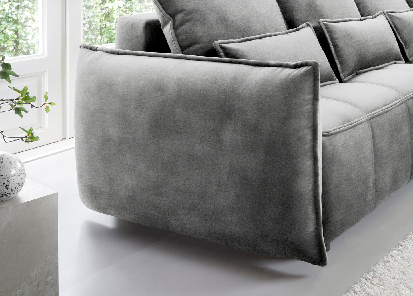 Detal zaokrąglonego podłokietnika i tylnej części narożnika Campo w układzie prawym, tapicerka Nube 04 szary na tle okna.