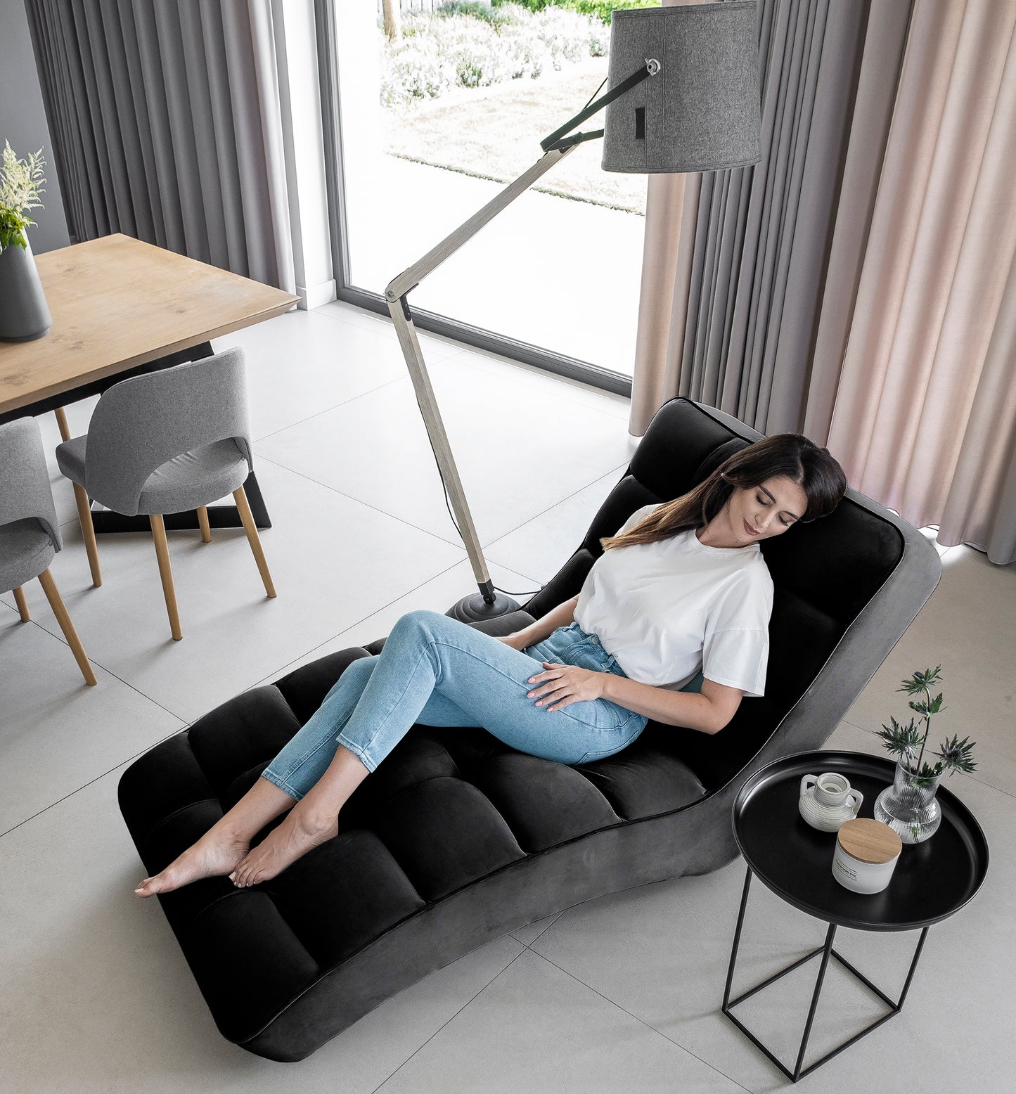 Kobieta relaksująca się na szezlongu Loren Loco 10 czarny w stylowym salonie z jasnymi zasłonami i nowoczesną lampą.
