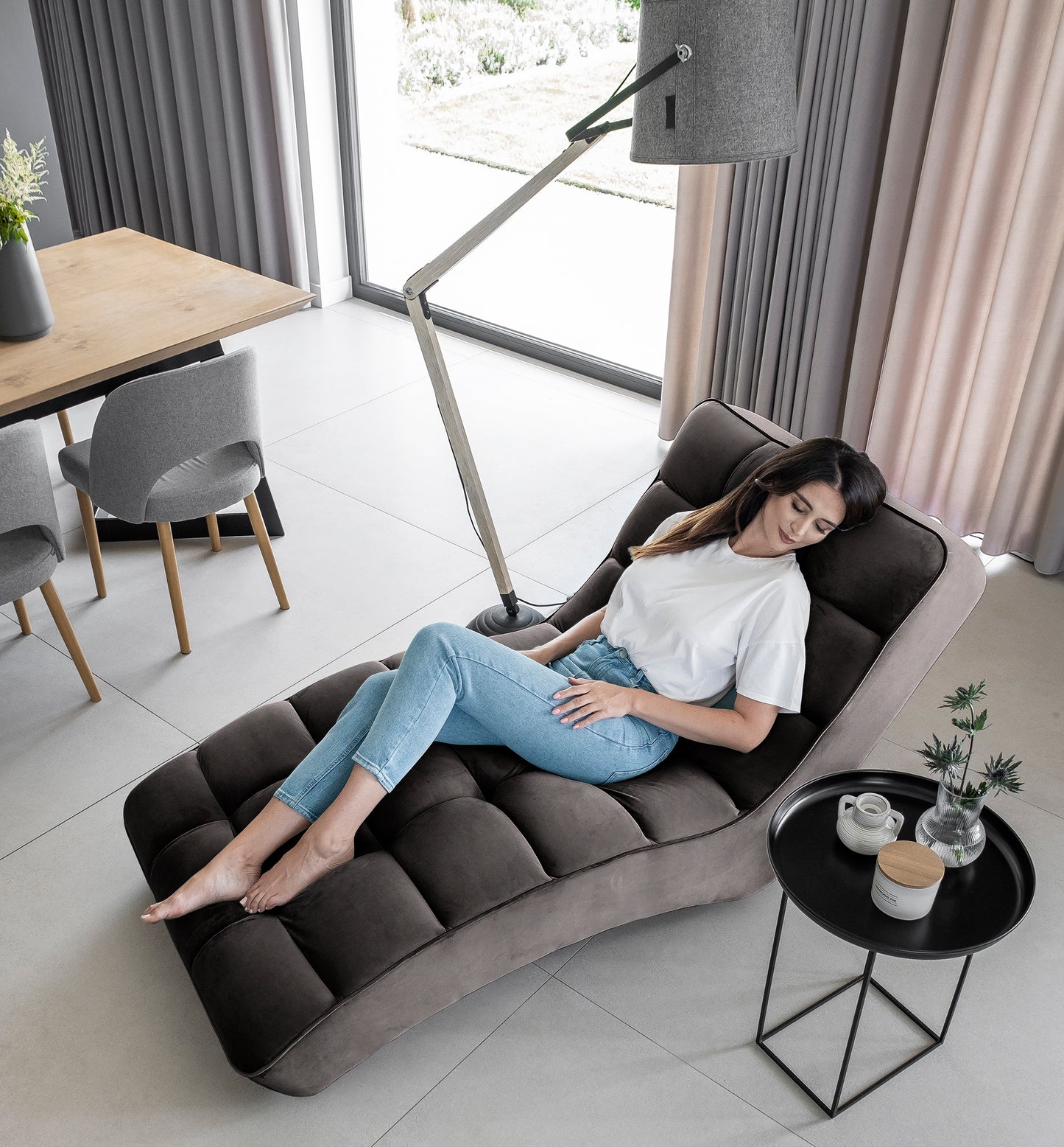 Kobieta w białej bluzce i dżinsach odpoczywa na ciemnoszarym szezlongu Loren w eleganckim salonie z dużym oknem.
