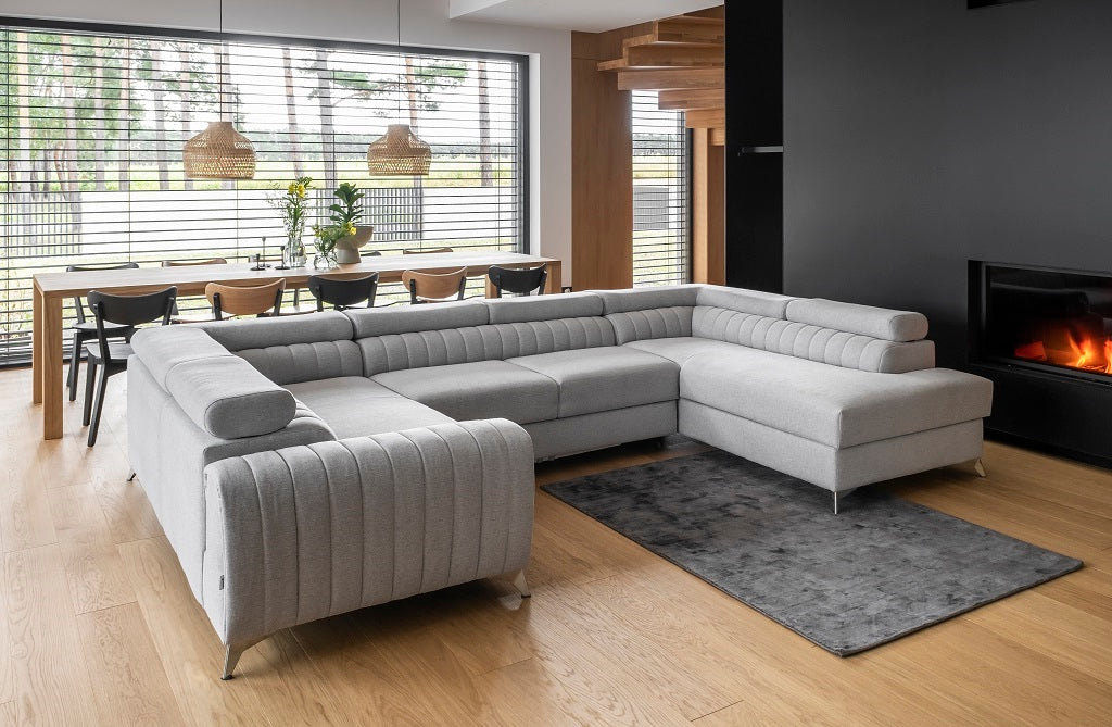 Przestronny salon z narożnikiem Louis w kolorze Borneo 04 szary w układzie prawym, ustawionym przy kominku na drewnianej podłodze, z dużym oknem i jadalnią w tle.