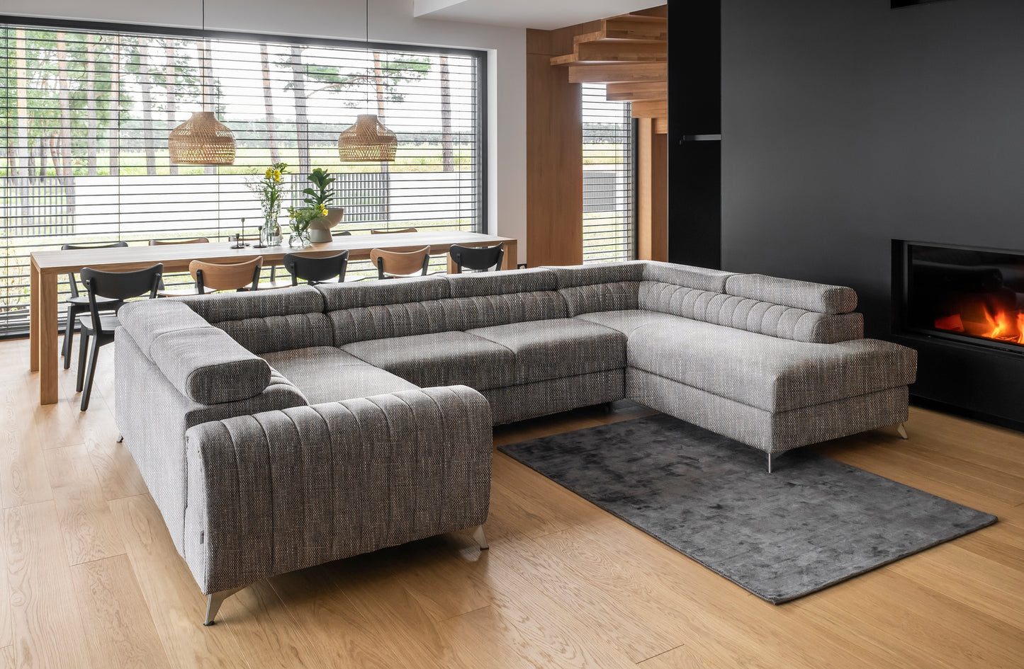 Przestronny salon z narożnikiem Louis w kolorze Marte 130 szary w układzie prawym, ustawionym przy kominku na drewnianej podłodze, z dużym oknem i jadalnią w tle.