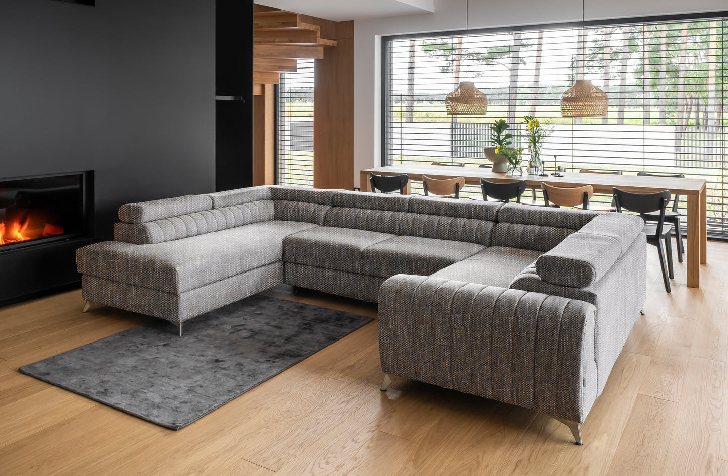 Przestronny salon z narożnikiem Louis w kolorze Marte 130 szary w układzie lewym, ustawionym przy kominku na drewnianej podłodze, z dużym oknem i jadalnią w tle.
