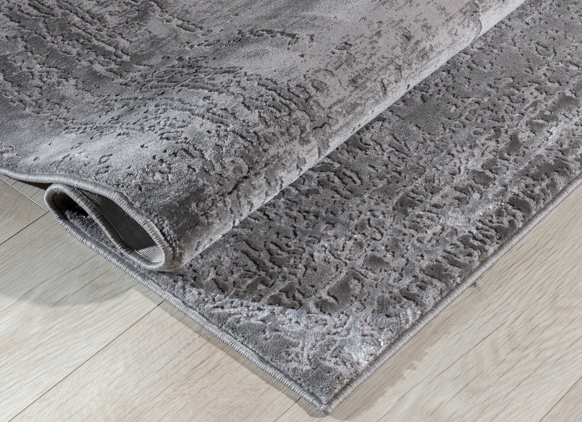 Dywan Codrila Grey 160x220 cm częściowo zwinięty, prezentujący grubość materiału i wysoką jakość wykonania.