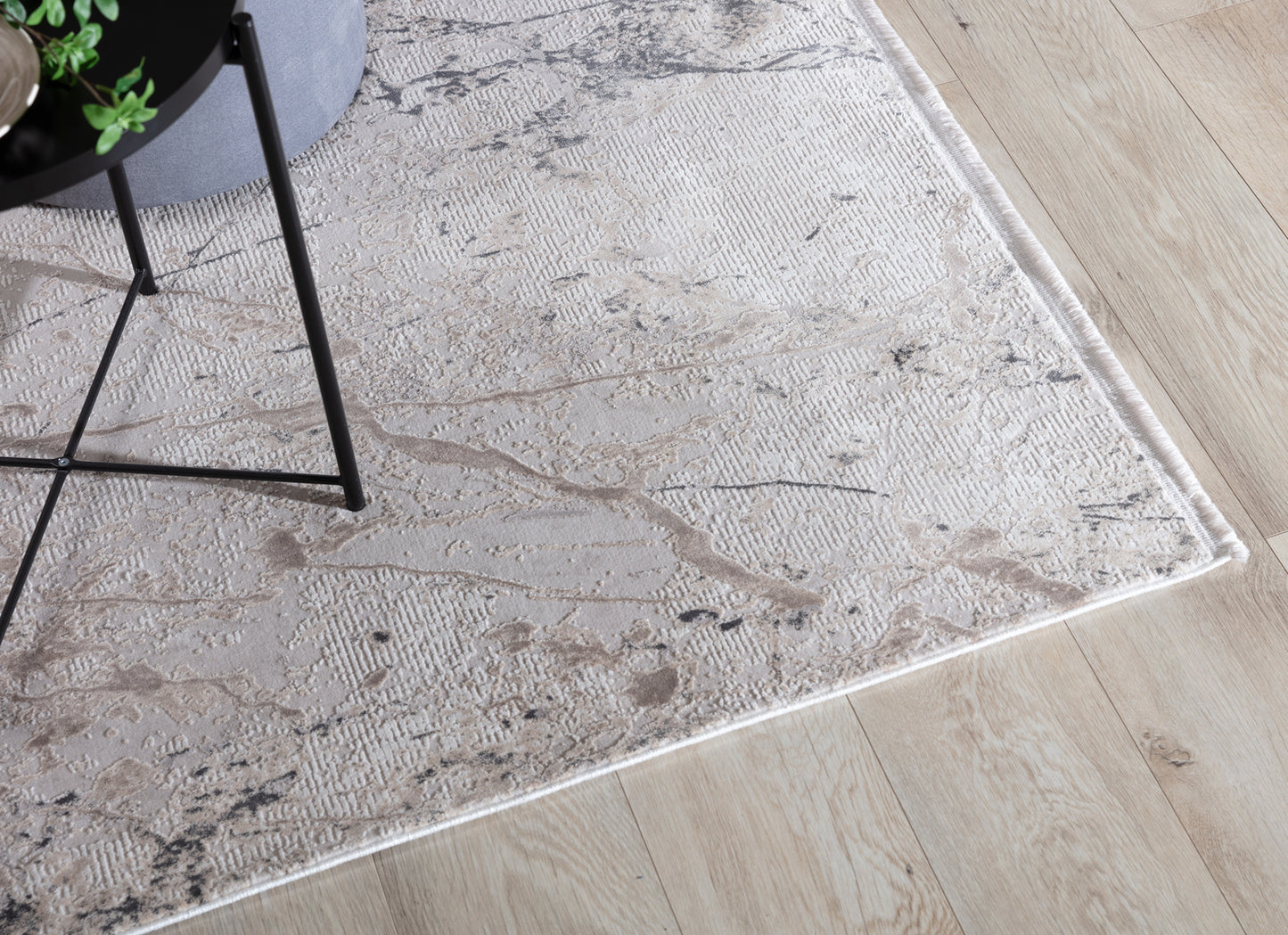 Zbliżenie na róg dywanu Collymore Beige 160x230 cm z marmurowym motywem, położonego na drewnianej podłodze obok stolika z czarnymi nogami.