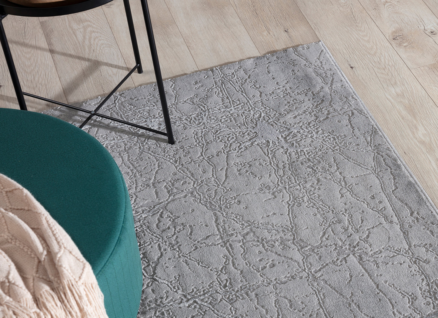 Szary dywan Siggi 120x180 cm z delikatnym, nieregularnym wzorem, ułożony na jasnej podłodze obok zielonego pufa i czarnego stolika.