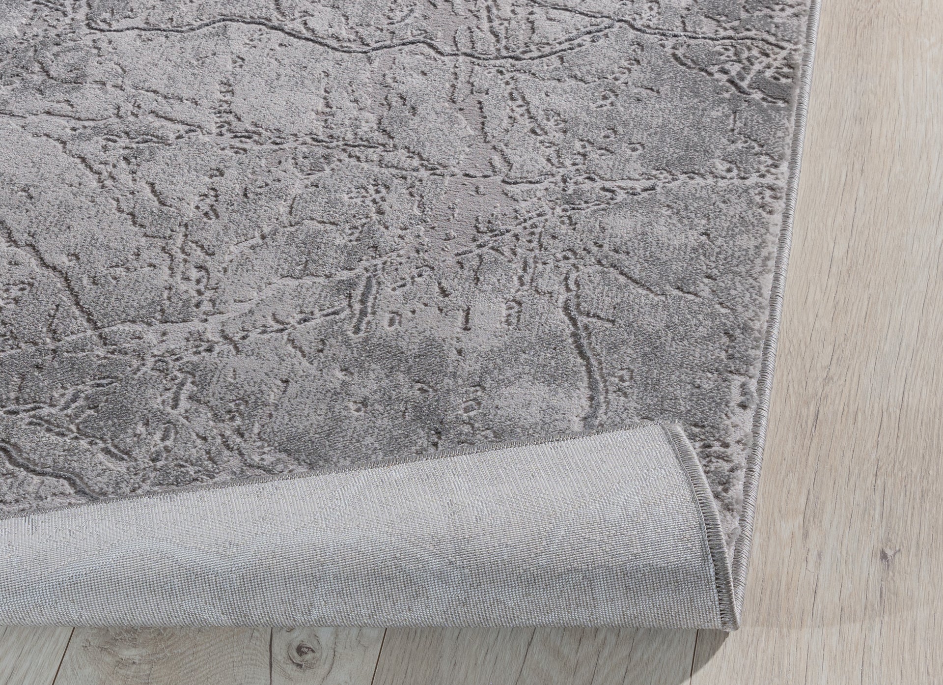 Dywan Siggi Grey 120x180 cm z lekko podwiniętą krawędzią – prezentacja elastyczności i spodniej warstwy tkaniny.