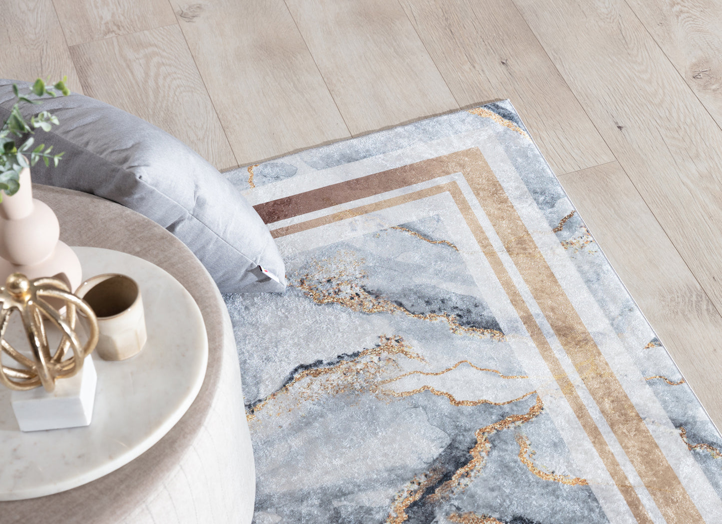 Dywan Squitta Grey 160x220 cm z marmurowym motywem i złotymi akcentami, ułożony na jasnej podłodze obok beżowego pufa i szarej poduszki.
