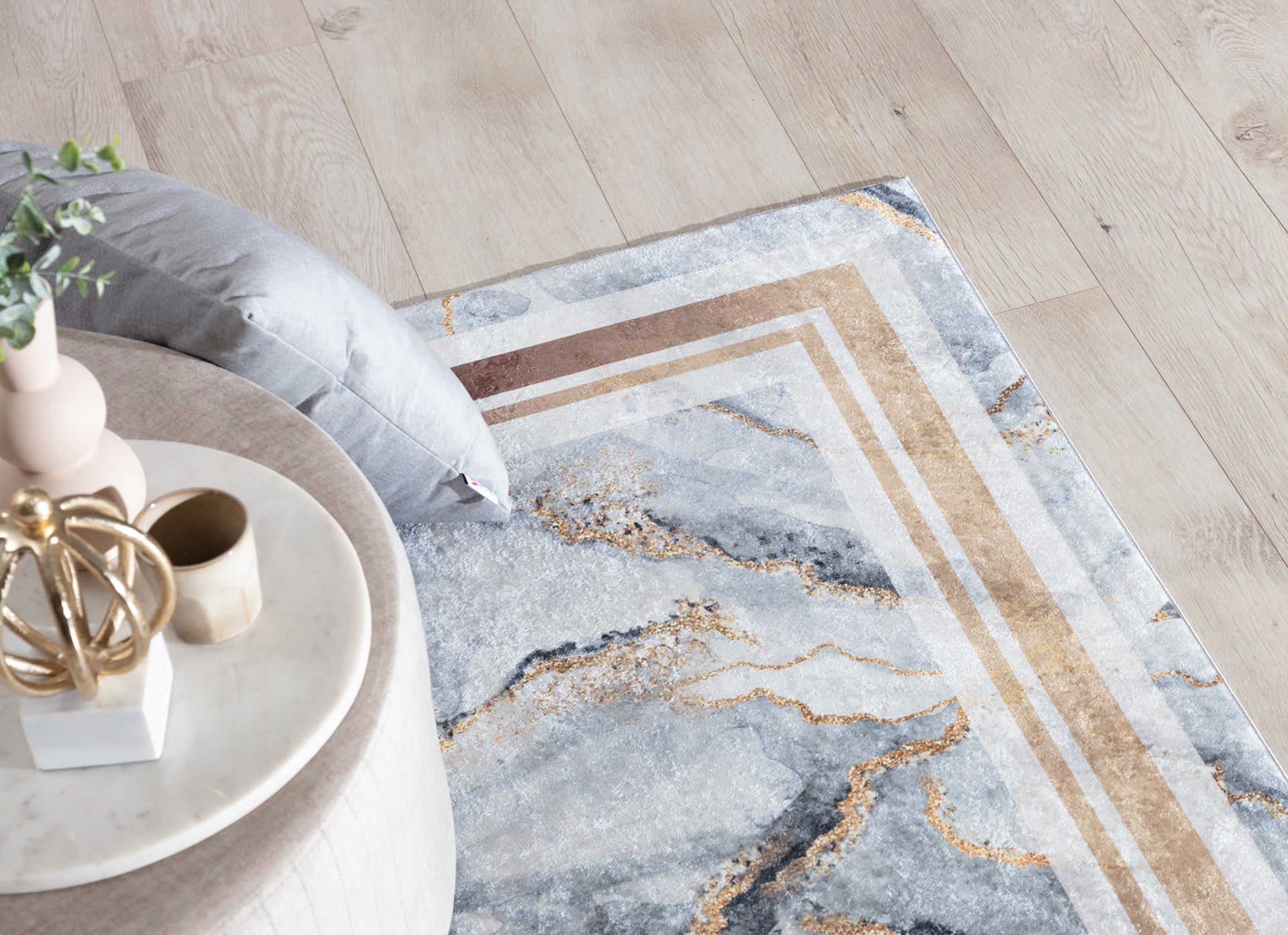 Dywan Squitta Grey 160x220 cm z marmurowym motywem i złotymi akcentami, ułożony na jasnej podłodze obok beżowego pufa i szarej poduszki.
