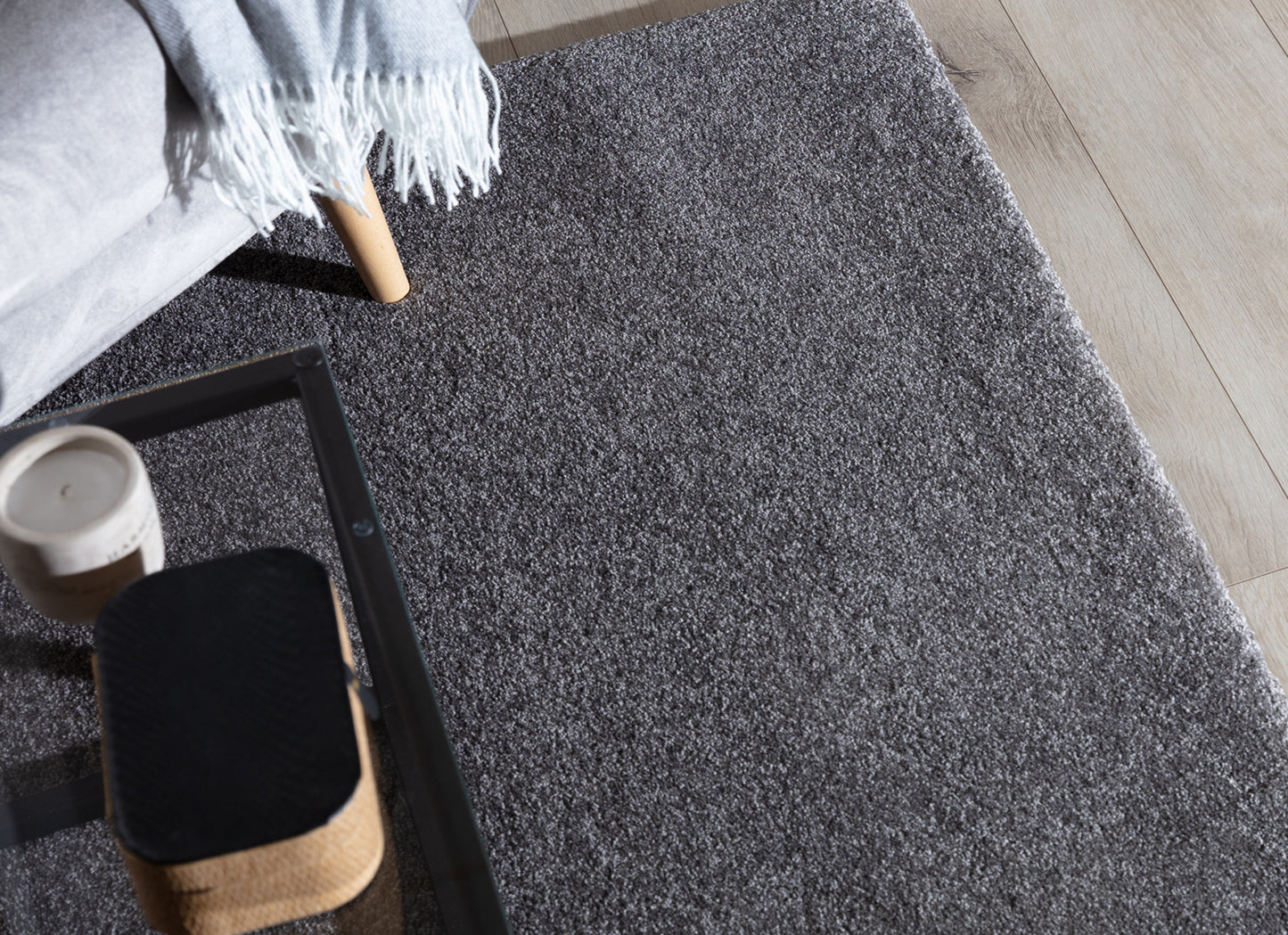 Szary dywan Verlice 80x150 cm ułożony przy sofie z kocem i stolikiem kawowym – widoczne miękkie runo i elegancka faktura.