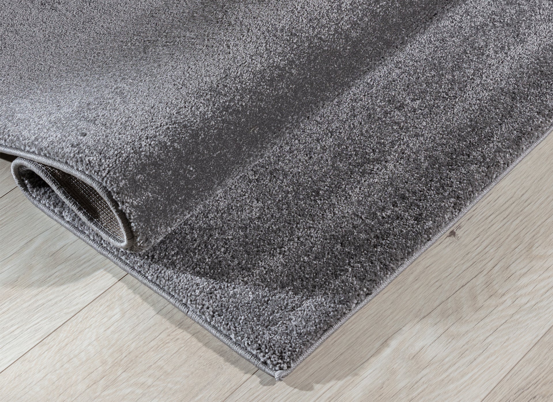 Dywan Verlice Grey 80x150 cm częściowo zwinięty – widoczna grubość i miękka, puszysta powierzchnia.