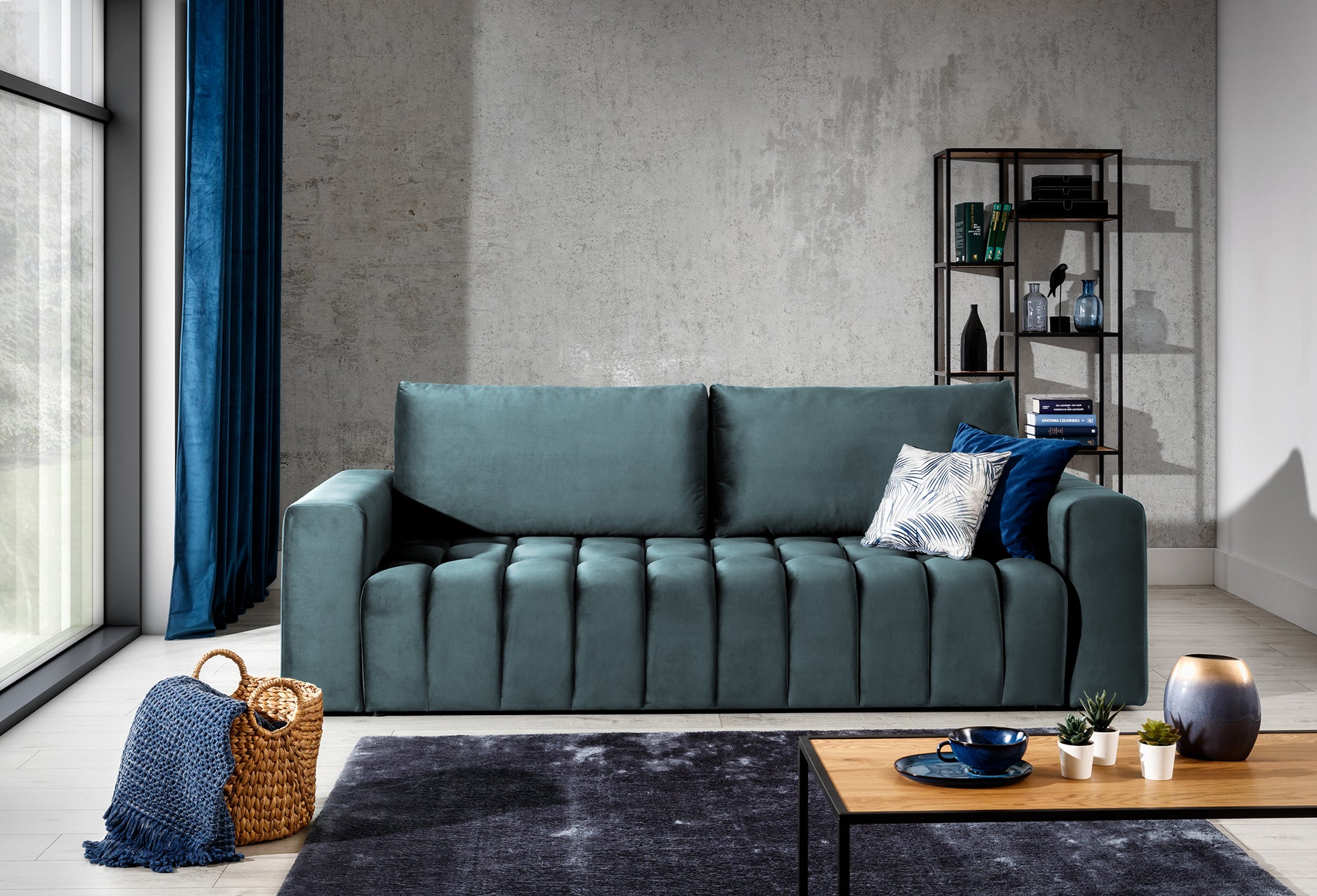 Sofa Lazaro Monolith 76 jasnoniebieska w aranżacji salonu: sofa ustawiona na tle betonowej ściany obok dużego okna z granatowymi zasłonami, uzupełniona metalowym regałem, wiklinowym koszem z kocem i stolikiem kawowym, podkreślając nowoczesny charakter wnętrza.