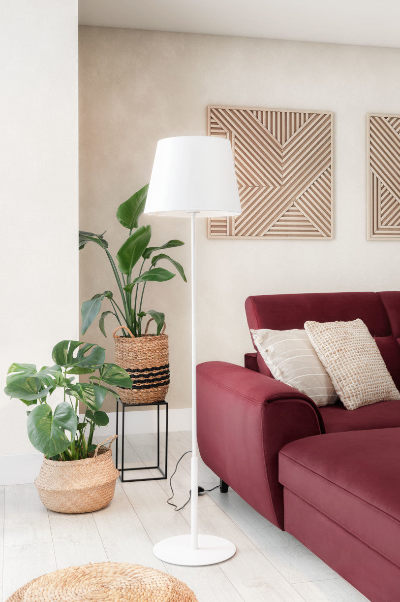 Lampa Vespillo 1a ustawiona obok czerwonej kanapy i donicy z monsterą w jasnym salonie z drewnianą podłogą i geometrycznymi dekoracjami ściennymi.