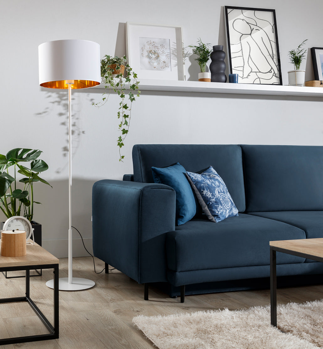 Lampa stojąca Vespillo w aranżacji salonu: obok granatowej sofy i drewnianego stolika z zegarem i rośliną, z półką z dekoracjami w tle.