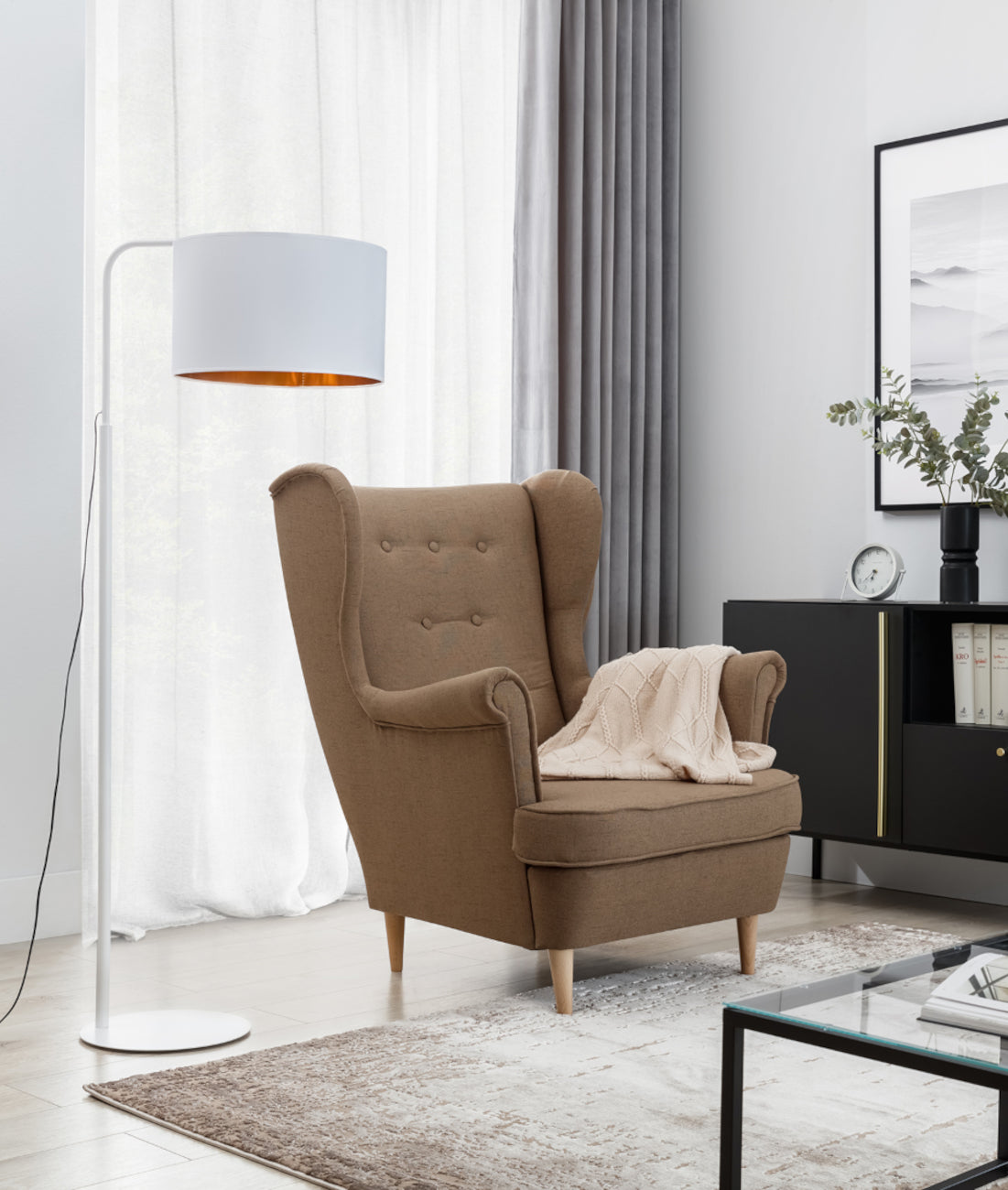 Lampa stojąca Vespillo 1 obok brązowego fotela uszaka w jasnym salonie z dużym oknem i białą zasłoną, ustawiona przy niskim stoliku z czarną metalową ramą.