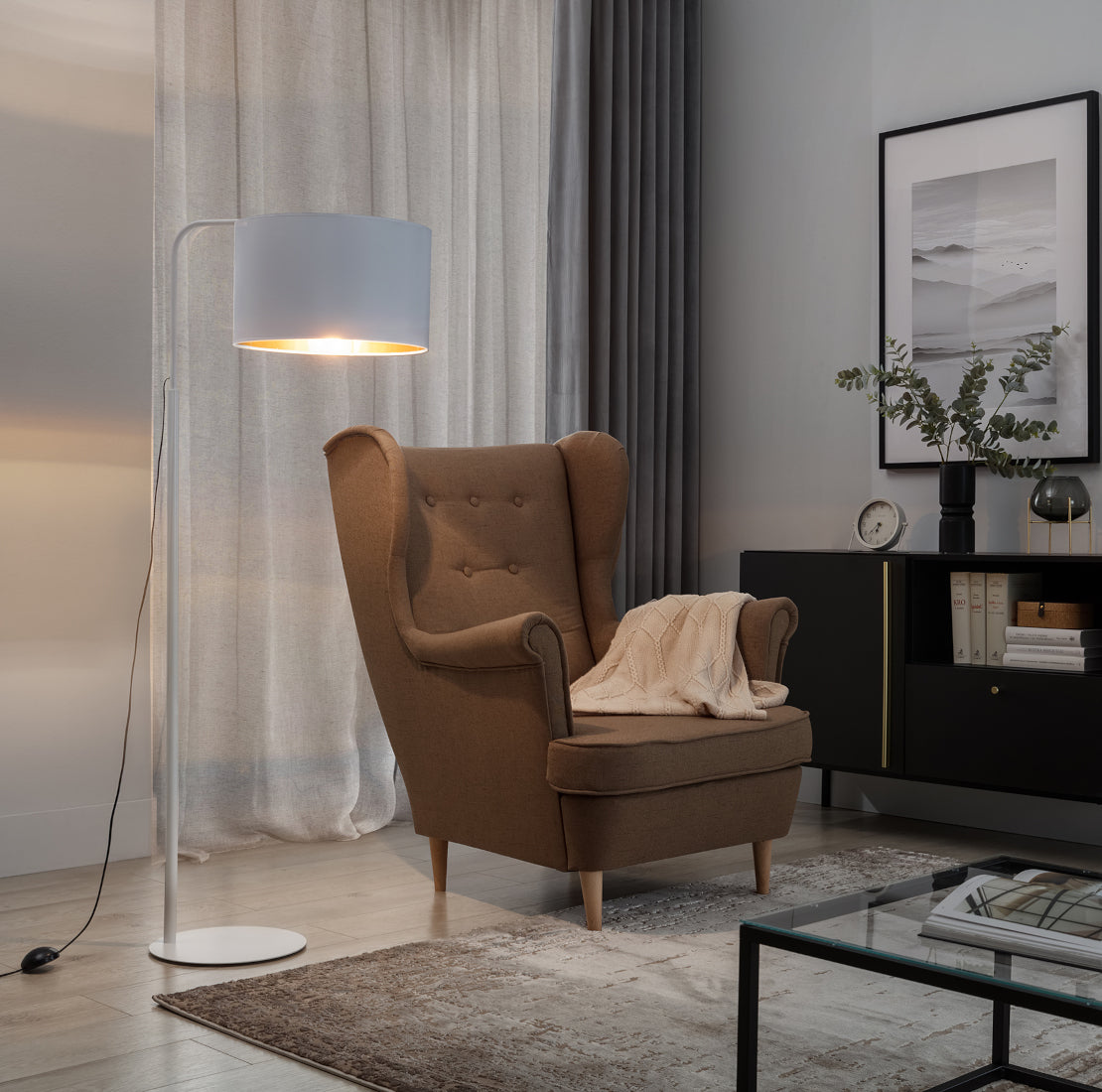 Włączona lampa stojąca Vespillo 1 rzucająca ciepłe, złote światło na fotel uszak i przylegającą ścianę w przytulnej, wieczornej aranżacji salonu.