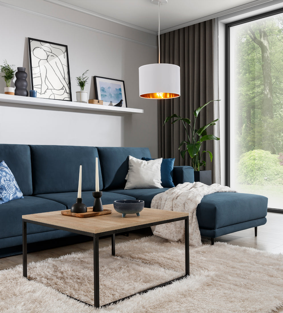 Lampa wisząca Vespillo w aranżacji salonu: nad stolikiem kawowym i granatową sofą przy dużym oknie, emitująca ciepłe światło ze złotego wnętrza abażura.