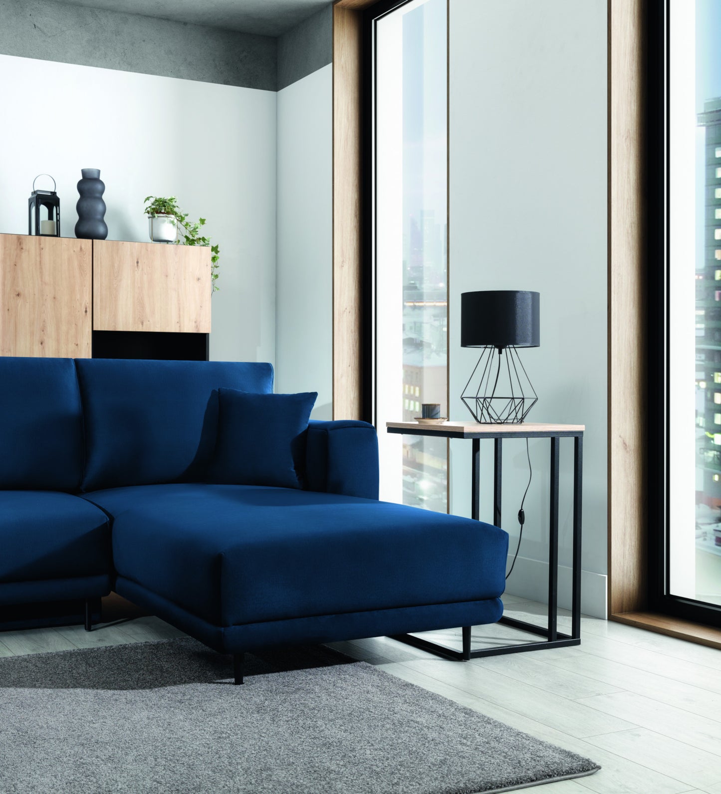 Lampka nocna Benigne 25x25 cm – aranżacja w salonie – lampka stołowa z czarnym abażurem ustawiona na stoliku pomocniczym obok granatowej sofy przy dużym oknie.