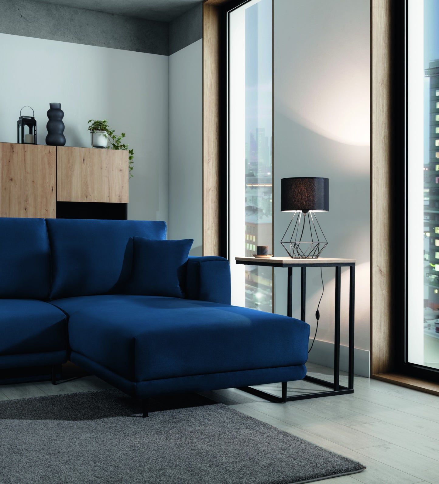 Lampka nocna Benigne 25x25 cm – aranżacja we wnętrzu z oświetleniem – lampka nocna z zapalonym światłem ustawiona na stoliku przy sofie w nowoczesnym salonie z widokiem na miasto.