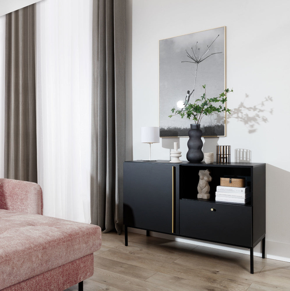 Lampka nocna Vespillo w kolorze biało-złotym ustawiona na czarnym kredensie obok różowej sofy, przy ozdobnych ramkach na ścianie i roślinie doniczkowej.