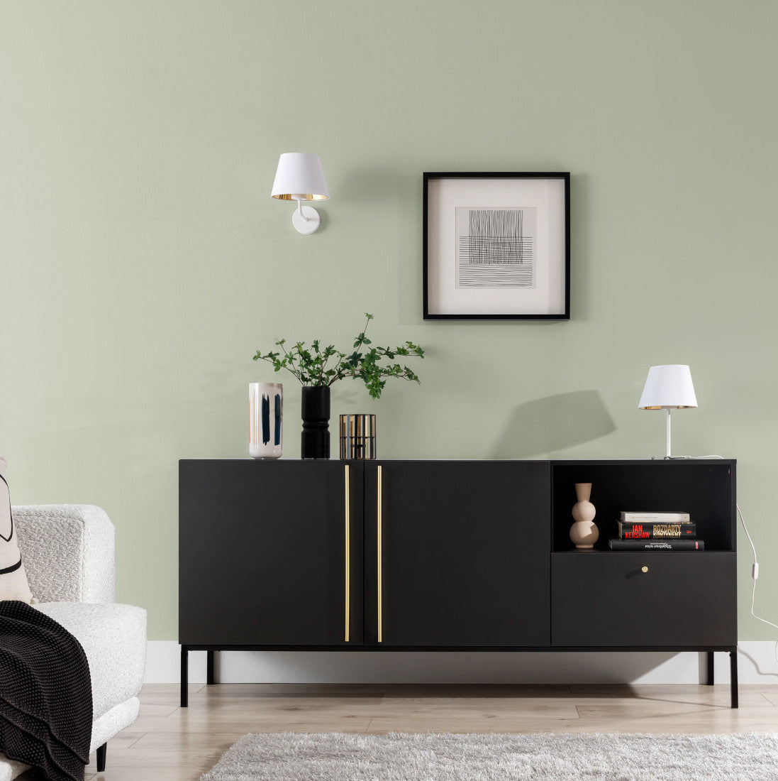 Biała lampa nocna Vespillo 1a ustawiona na czarnej, niskiej komodzie przy jasnej, pastelowej ścianie, obok ramki z minimalistycznym obrazem i dekoracyjnych wazonów; przy lampce stoi biały narożnik i duże okno z widokiem na zieleń.