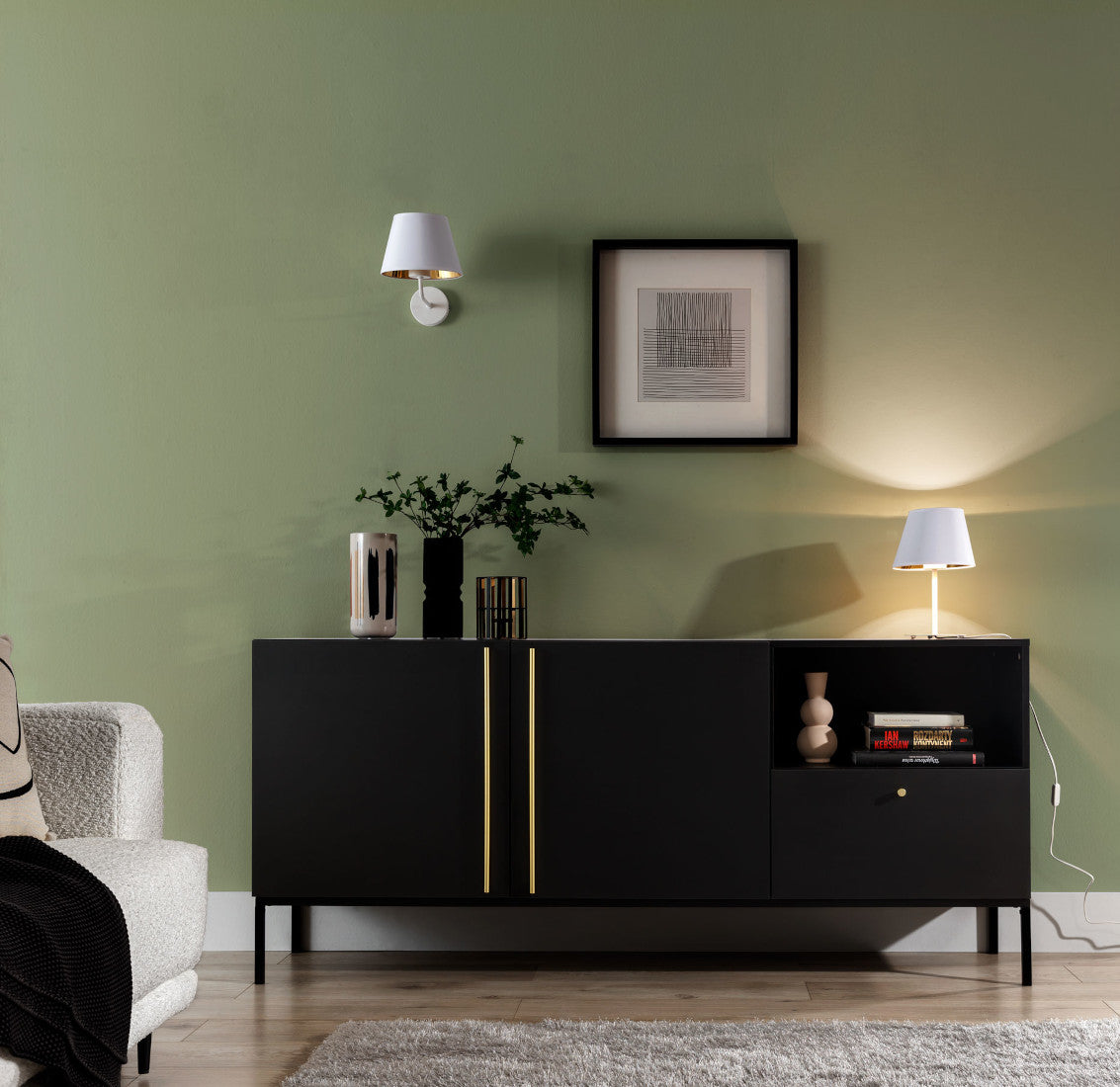 Lampa nocna Vespillo 1a włączona po zmroku, emitująca ciepłe światło, umieszczona na czarnej komodzie na tle przytulnego salonu z zieloną ścianą i dekoracjami.