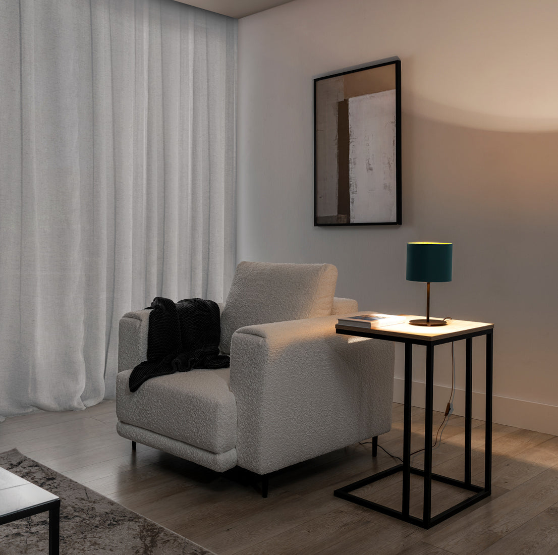 Włączona lampka Vespillo rzucająca ciepłe, złote światło na stolik i fotel w przytulnej, wieczornej aranżacji salonu z półprzezroczystą zasłoną i dekoracyjną grafiką na ścianie.