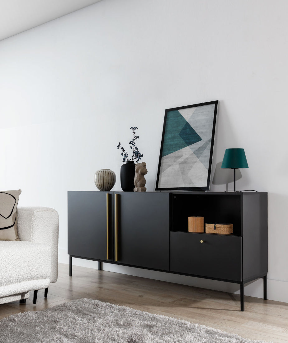 Lampka Vespillo 2a ustawiona na czarnym bocznym stoliku obok beżowego fotela, we wnętrzu z białą ścianą, grafiką w ramie i czarną komodą, tworząca elegancki kontrast.