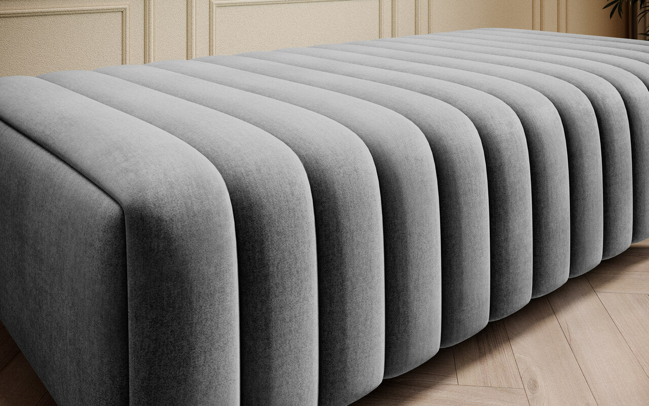 Tapicerowana pufa ławka Laurence w szarej tkaninie Nube 04, prezentowana jako praktyczny mebel pomocniczy z prążkowanym siedziskiem do nowoczesnych i eleganckich wnętrz.