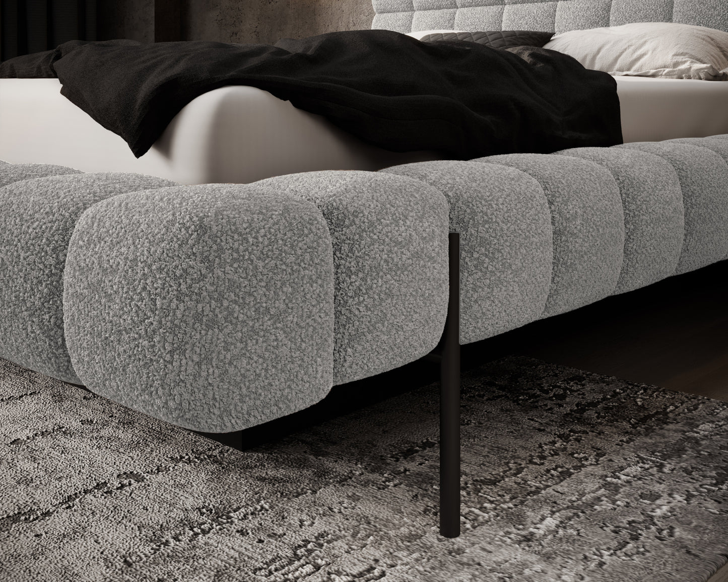Zbliżenie na dolną ramę łóżka Magentta tapicerowaną tkaniną Royal 04 w jasnoszarym odcieniu, z widoczną czarną metalową nogą na tle dywanu.