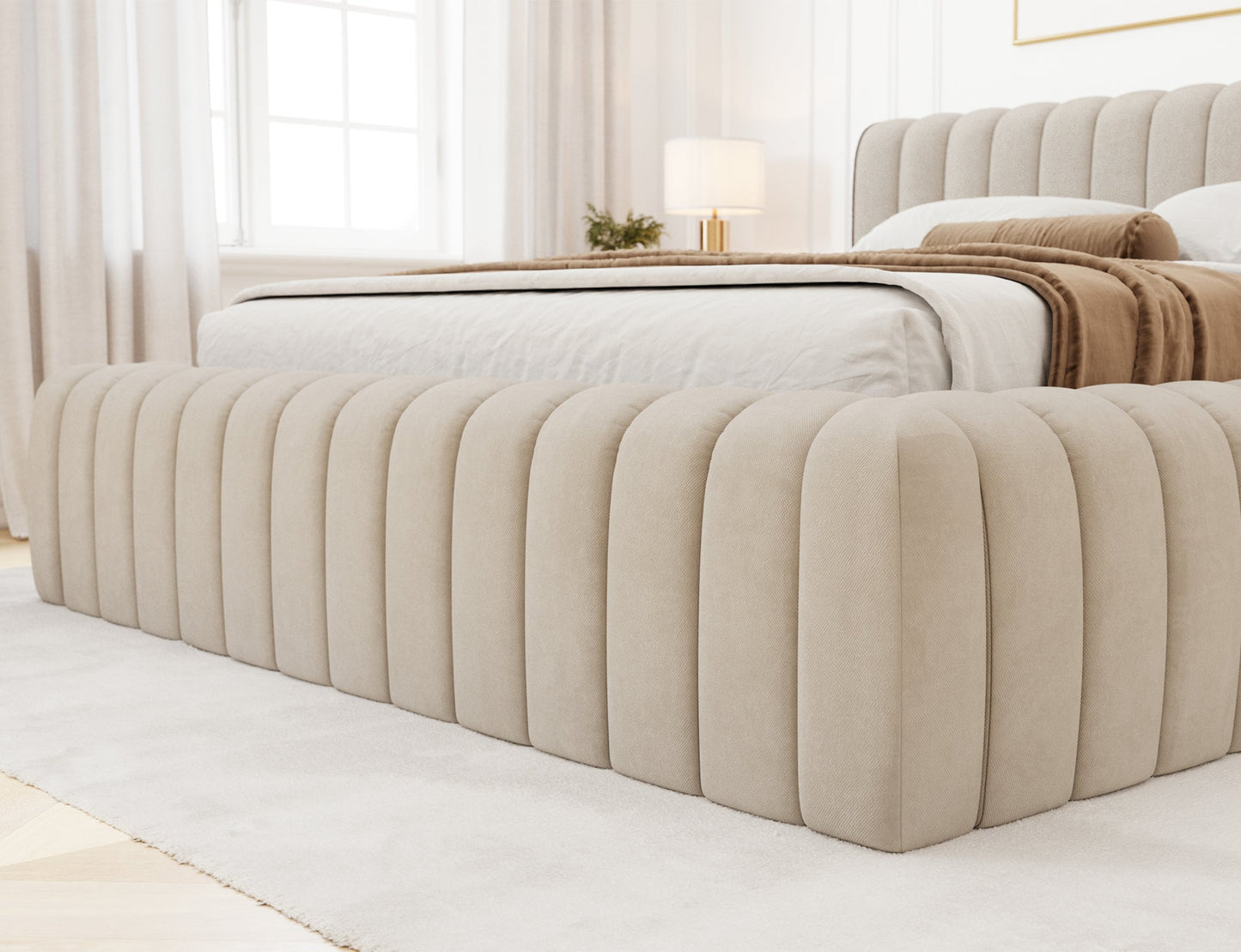 Detal narożnika łóżka Carilla tapicerowanego tkaniną Sola 18 w kolorze beżowym, ustawionego w jasnej, eleganckiej sypialni.