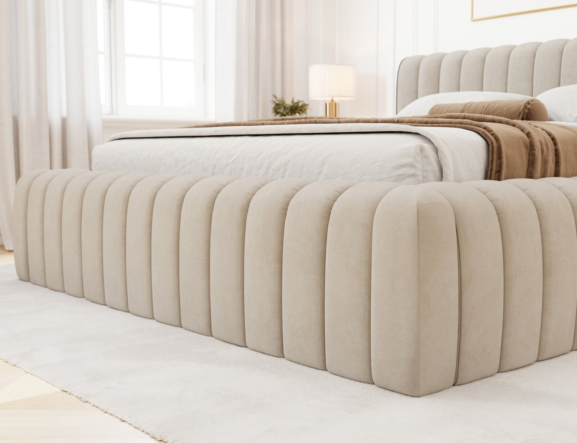 Detal narożnika łóżka Carilla tapicerowanego tkaniną Sola 18 w kolorze beżowym, ustawionego w jasnej, eleganckiej sypialni.