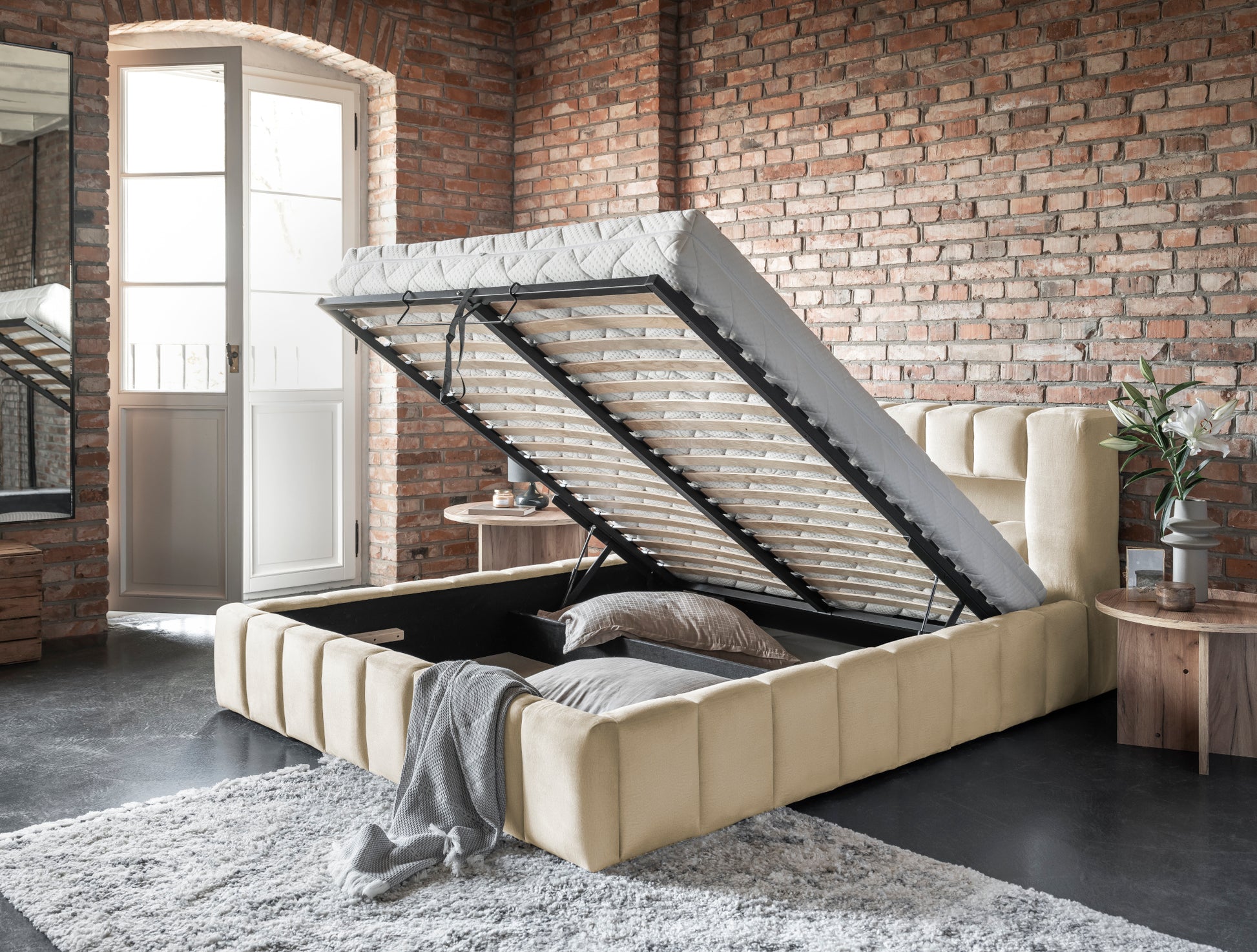 Beżowe łóżko Lamica z podniesionym stelażem i pojemnikiem – aranżacja w lofcie z ceglaną ścianą.