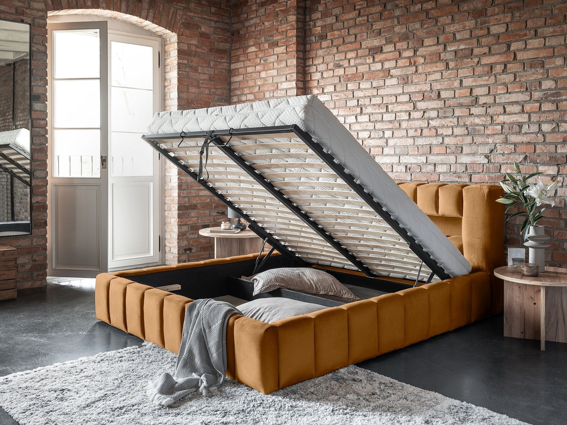 Łóżko Lamica Monolith 48 miodowy (140 × 200 cm) z uniesionym stelażem odsłaniającym pojemny schowek na pościel, na tle surowej, ceglastej ściany i stolika z dekoracją.