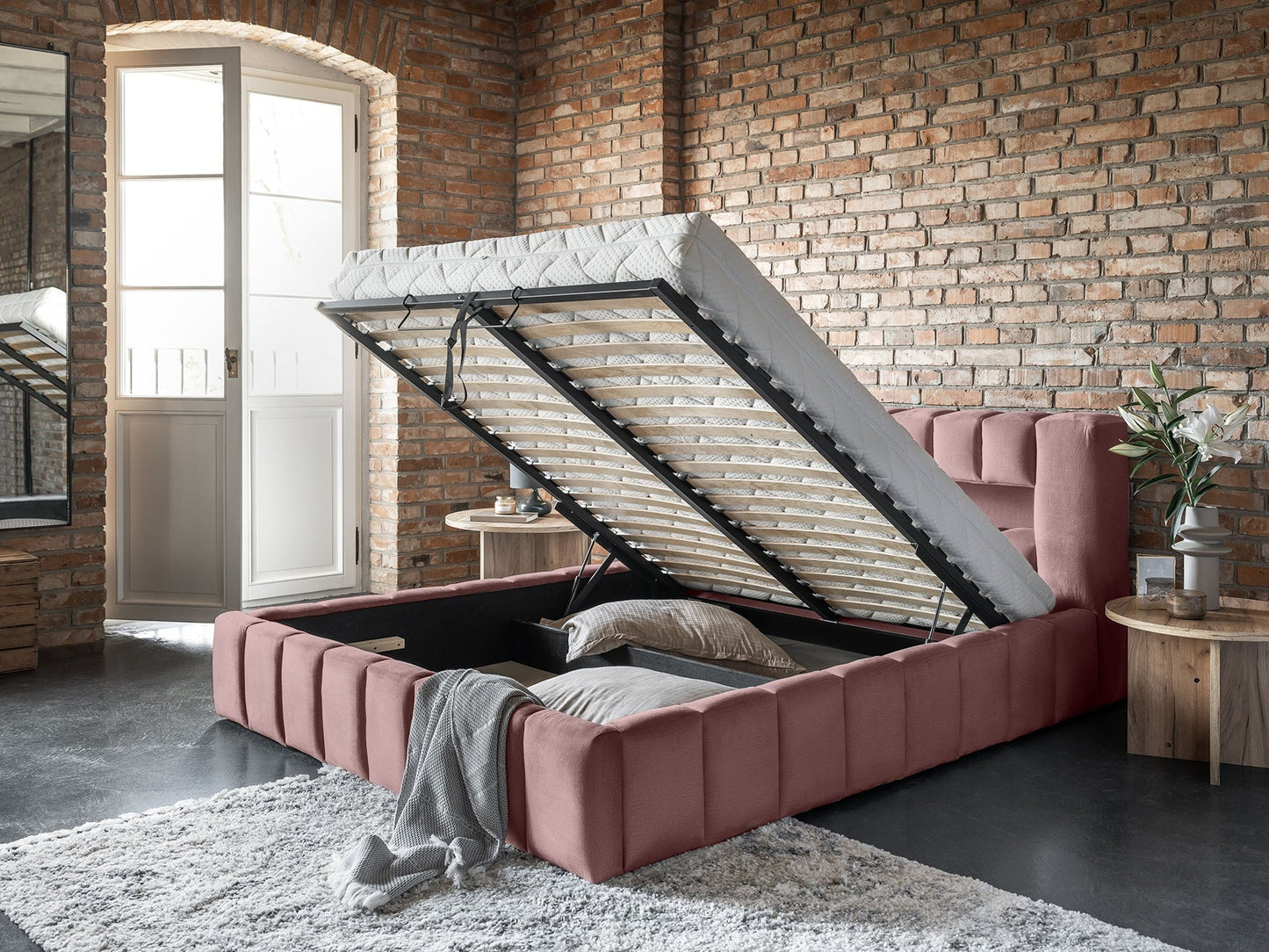Zbliżenie na łóżko Lamica Monolith 63 różowy (140 × 200 cm) z uniesionym stelażem odsłaniającym obszerną, zamykaną przestrzeń do przechowywania na tle surowej, ceglastej ściany.