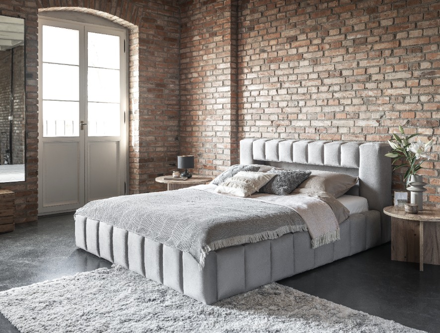 Łóżko Lamica Monolith 84 jasnoszary (140 × 200 cm) ustawione na tle surowej, ceglastej ściany w przestronnej sypialni loftowej, z dekoracyjną stolikową lampką i rośliną.